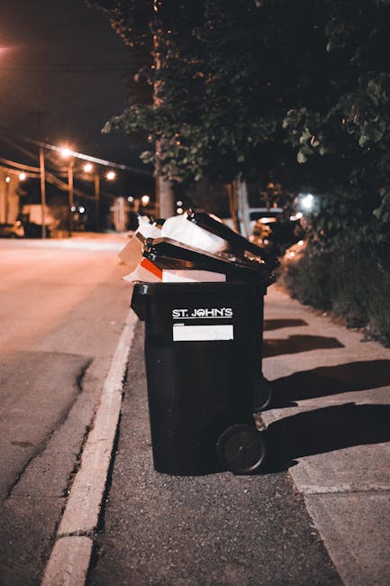 A man with dark hair wearing a black T-shirt is actively engaged in rubbish collection at an outdoor public space. He is bending forward, placing a large white plastic bag filled with waste into a mod