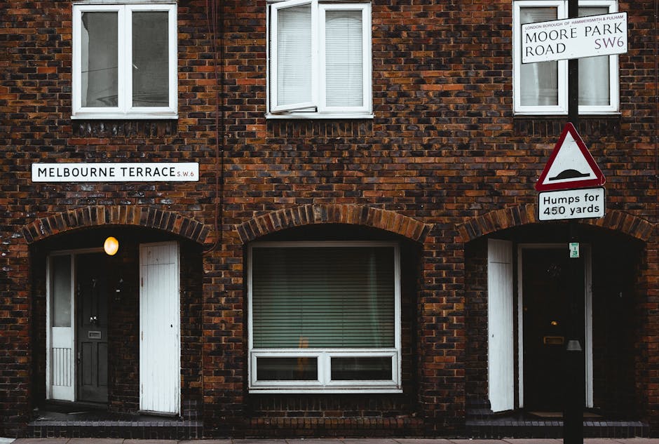 The image depicts a brick residential building with a façade featuring three white-framed windows; two are located on the upper floor, with one partially open, and one on the ground floor, with close