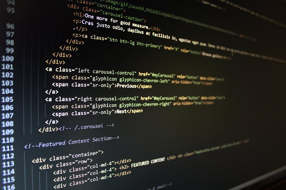 A close-up view of a person's hand using a black keyboard on a desk, with a computer monitor displaying lines of code in the background. The desk surface is light-colored, and nearby are a white mug, 