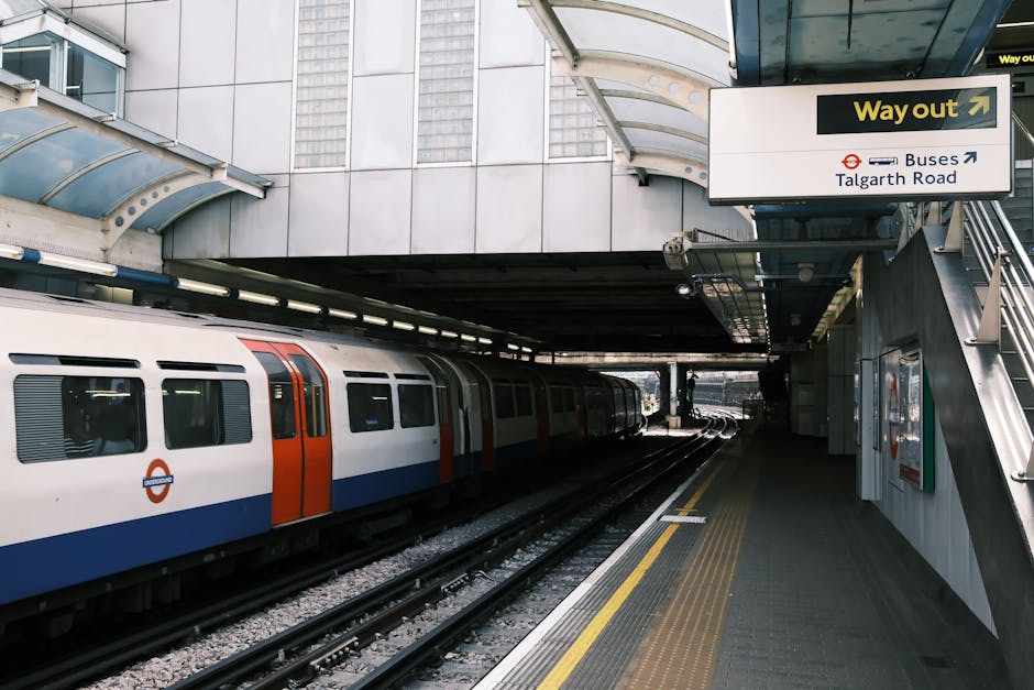 A modern London underground train with a white body, orange and blue accents, and the roundel logo, parked at a station platform beneath a curved glass and metal canopy. The platform features yellow t