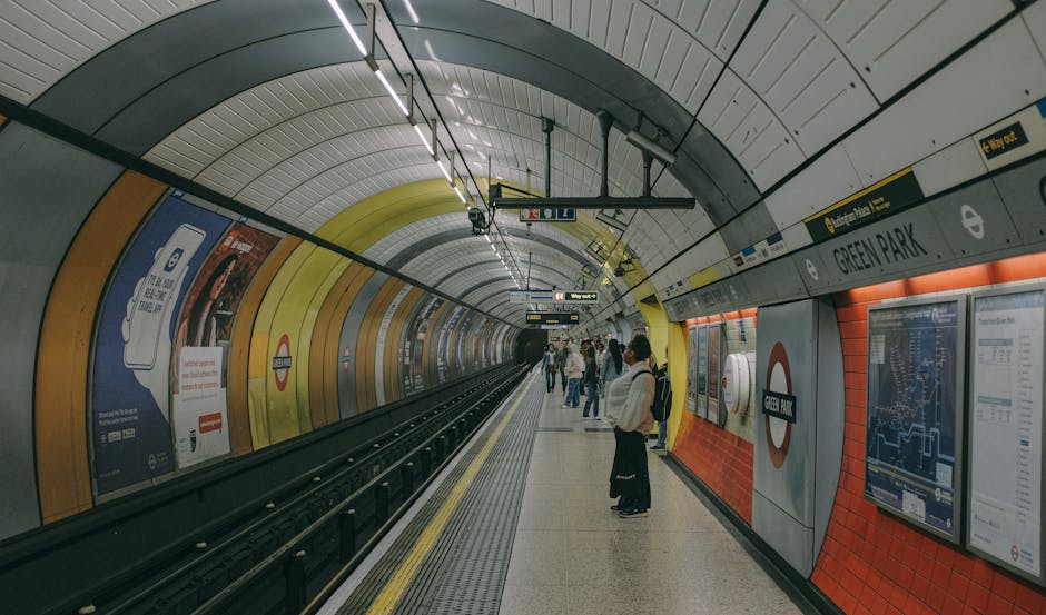 A modern London underground train with a white body, orange and blue accents, and the roundel logo, parked at a station platform beneath a curved glass and metal canopy. The platform features yellow t